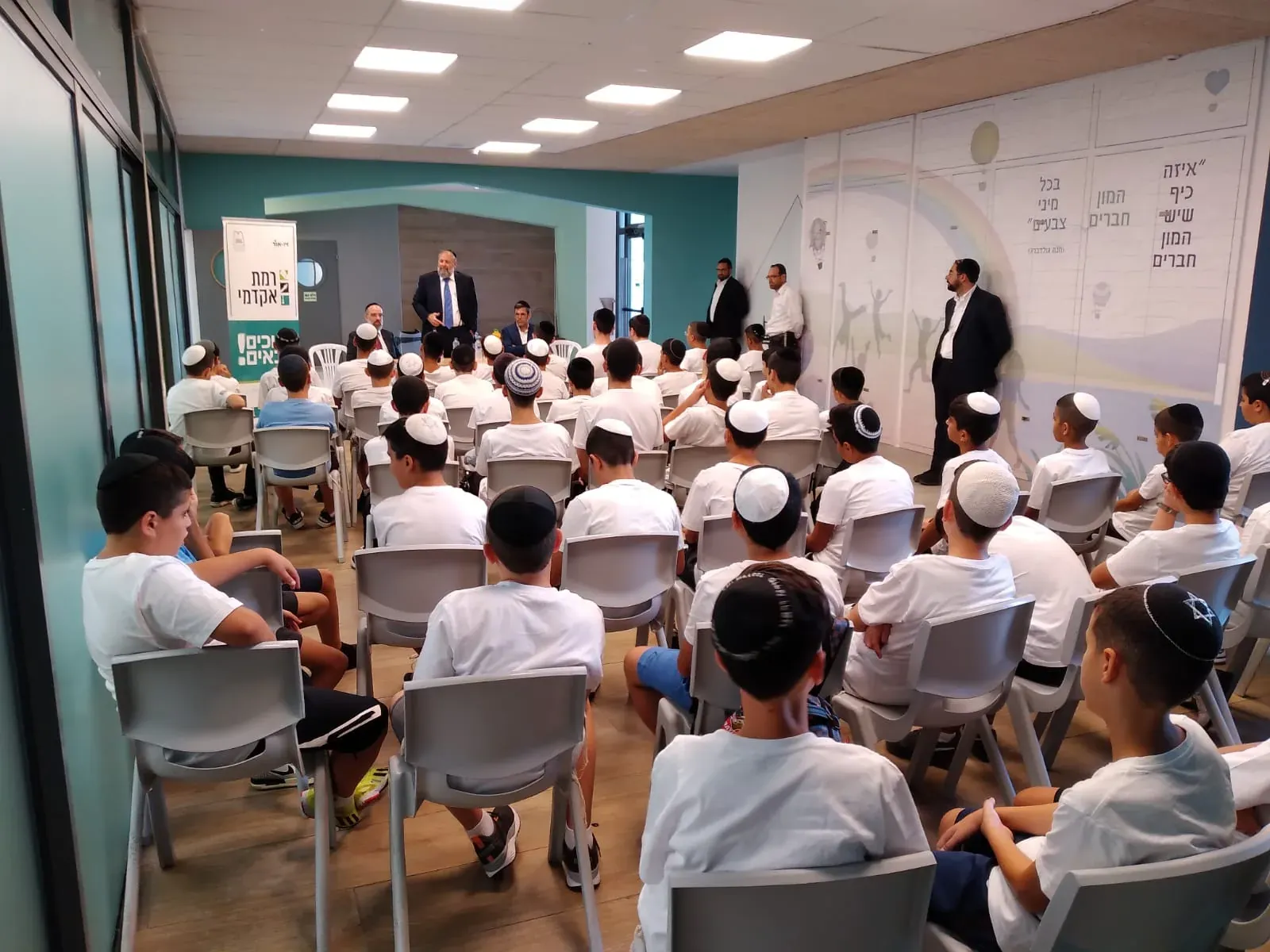 תלמידים יושבים באסיפה, צוות בית הספר מאחור